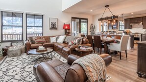 A spacious living room with a large brown leather sofa, a coffee table, and a dining area with chairs. There is a kitchen in the background with modern appliances and pendant lighting.