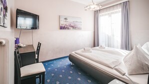A hotel room with a bed, a desk with a chair, a TV mounted on the wall, and a painting on the wall. - Hotel Kärntnerhof Velden by S4Y (Velden am Wörther See)