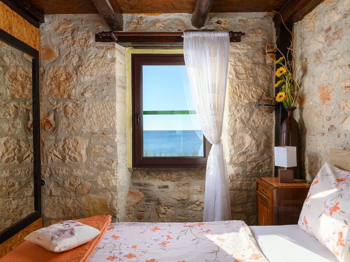A bedroom with stone walls, a wooden ceiling, a bed with floral bedding, a nightstand with a lamp, and a window with a view of the sea.