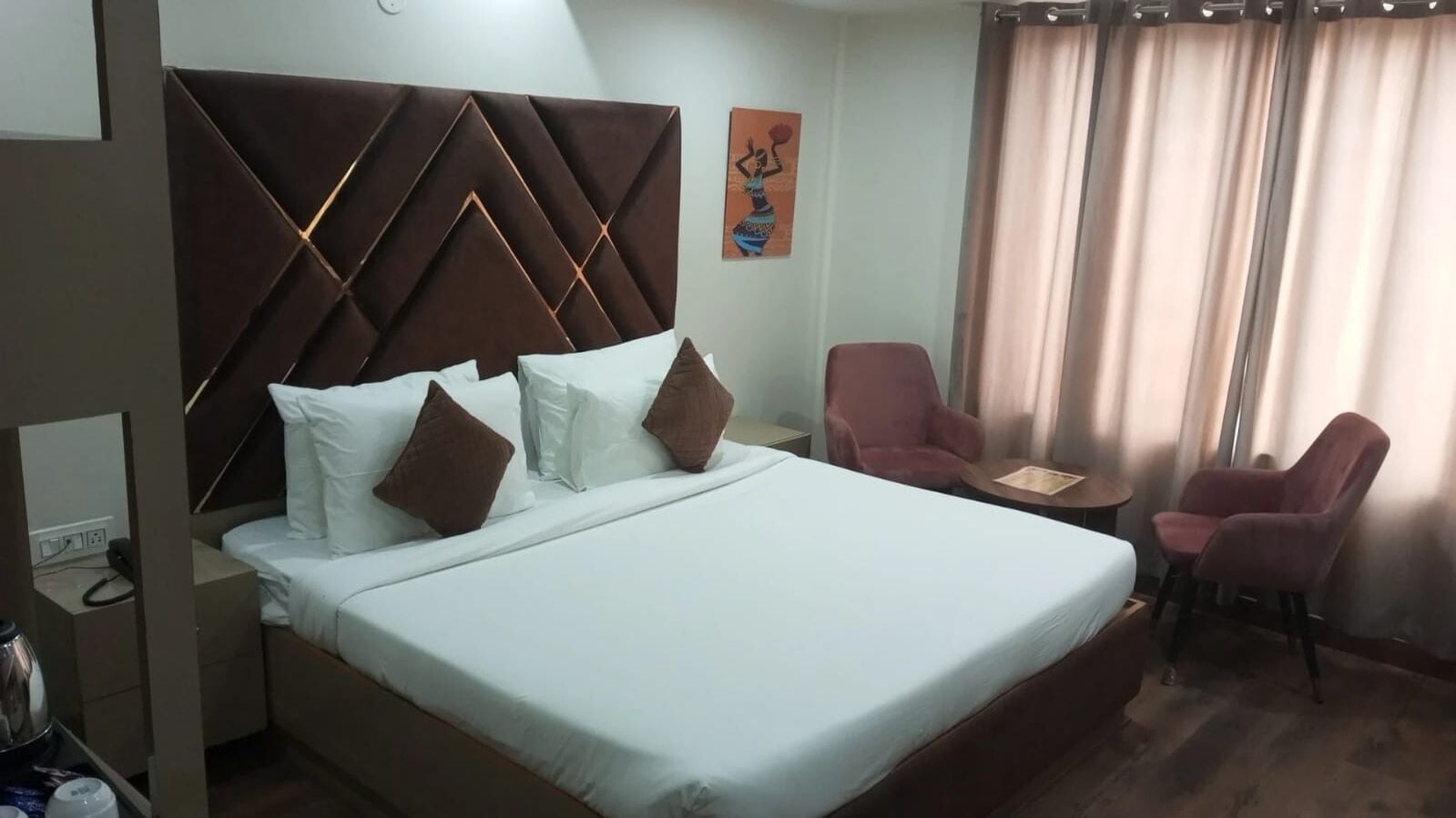 A hotel room with a large bed, a headboard with a geometric pattern, a small table, two chairs, and a painting on the wall.