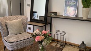 Une pièce comprenant un fauteuil à motifs, un miroir, une étagère avec un livre, un vase avec des fleurs et une plante en pot.