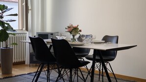 Un coin repas avec une table dressée pour quatre personnes, des chaises noires, une plante en pot et une vue sur un bâtiment par la fenêtre.