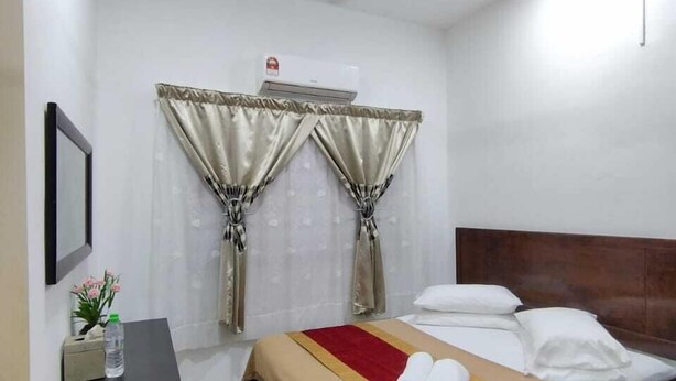 A hotel room with a bed, a bedside table with a bottle, a trash bin, a ceiling fan, and curtains.