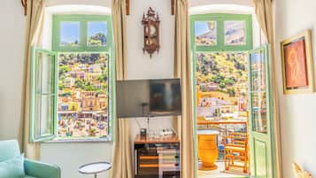Ein Zimmer mit einem großen Fernseher, einer Holzkommode und Blick auf eine farbenfrohe Stadt durch grün gerahmte Fenster.