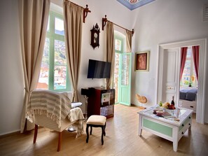 A room with a bed, a television, a table with fruit and wine, and a view of a town through the windows. - Athenas - Neoclassic Harbour View (Symi)
