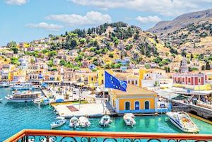 Un pueblo costero con edificios coloridos, un puerto deportivo con barcos y una colina cubierta de vegetación.