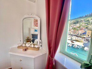 A room with a white dresser, a mirror, and a view of a coastal town through a window with a red curtain. - Athenas - Neoclassic Harbour View (Symi)