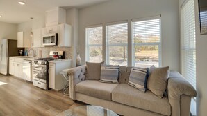 Una cocina moderna con armarios blancos, microondas integrado y estufa a gas. Hay un sofá color beige con cojines estampados frente a amplias ventanas.