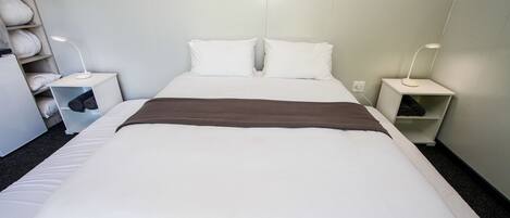 A neatly made bed with white linens and a brown throw blanket, two bedside tables with lamps, and a shelf with rolled towels.