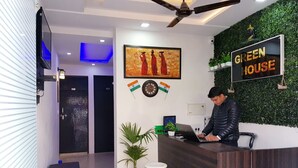 Uma área de recepção com um balcão, uma pessoa trabalhando em um laptop, uma placa na parede com a inscrição "GREEN HOUSE" e uma obra de arte emoldurada sobre o balcão.