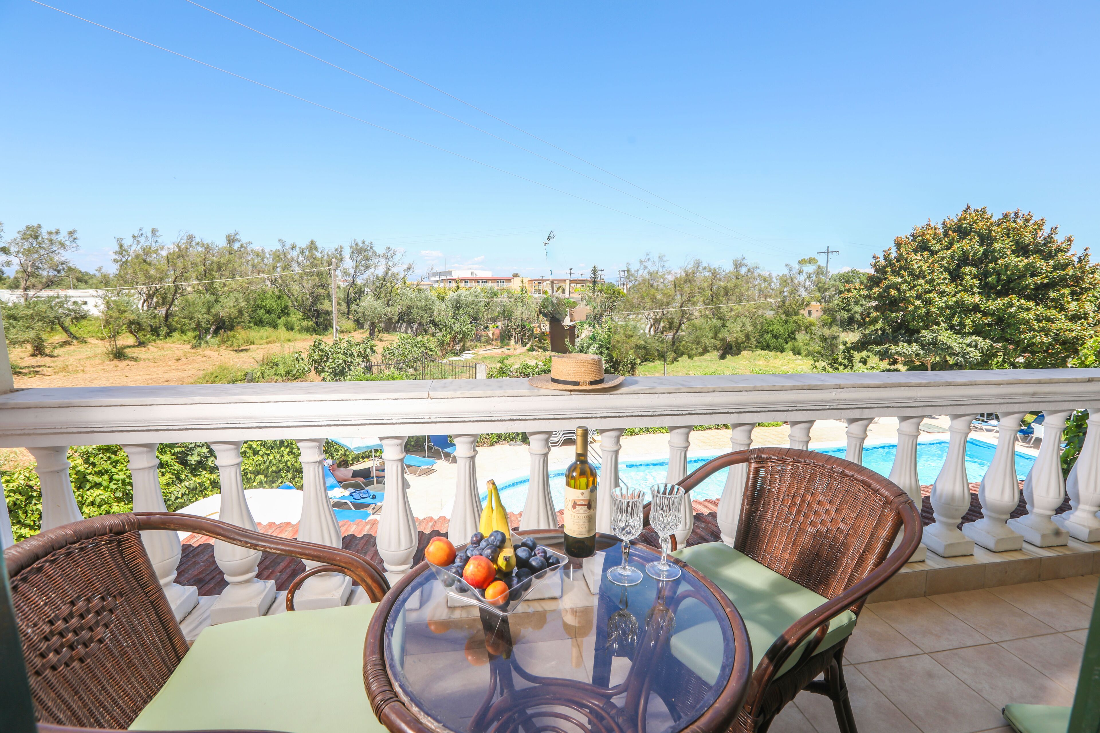 Un balcon avec des fauteuils en osier, une table garnie de fruits et de vin, et une vue sur une piscine et la verdure.
