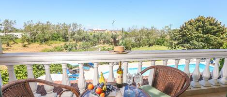A balcony with wicker chairs, a table with fruit and wine, and a view of a swimming pool and greenery.