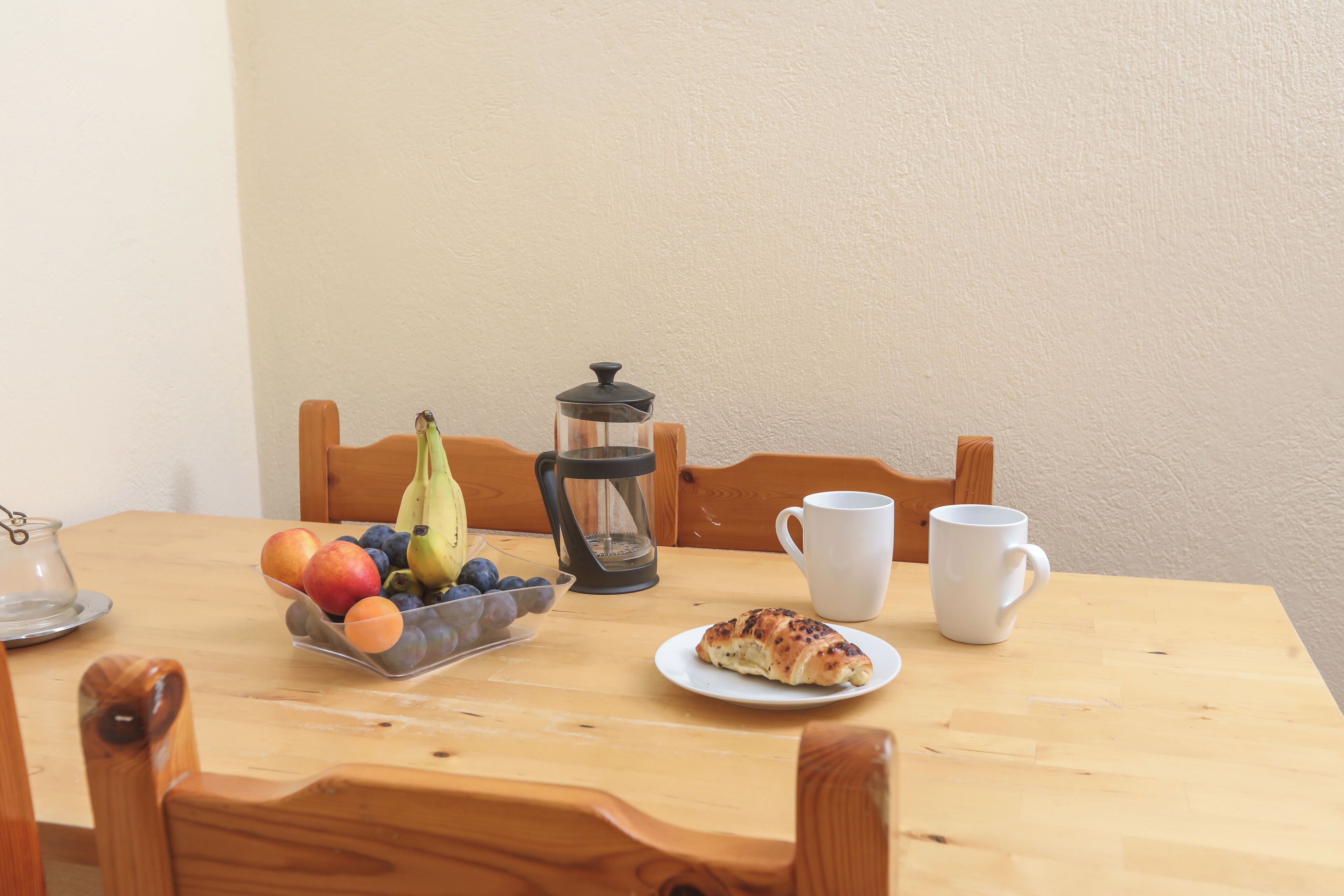 Una mesa de madera con un frutero, una cafetera, una taza y un plato con un croissant.