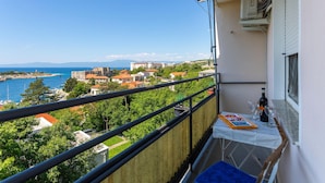 A balcony with a table set for two, overlooking a coastal town with buildings and a clear blue sky. - Lovely apartment in Kraljevica (Kraljevica)