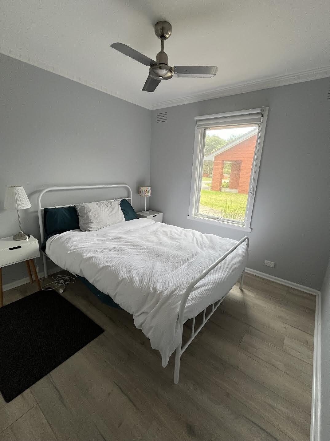 A bedroom with a single bed, a nightstand, a lamp, a ceiling fan, and a window with a view of a brick building.