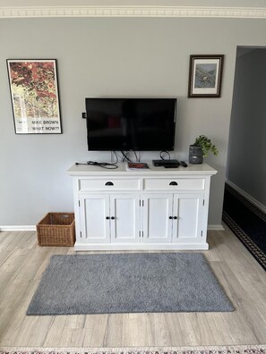 Un salon avec un téléviseur à écran plat fixé sur un meuble blanc, un tapis gris et deux tableaux encadrés accrochés au mur.