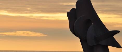 Une sculpture représentant une main à la forme courbée, placée sur fond de coucher de soleil sur l’océan.