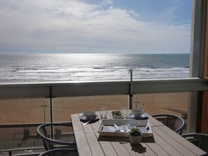 Une table de balcon avec un plateau, des bols, des verres et une cuillère, donnant sur une plage où l’on peut voir les vagues.