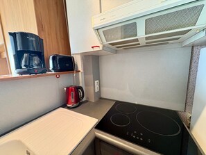 Eine kompakte Küchenzeile mit einer schwarzen Induktionskochplatte, einer weißen Arbeitsplatte, einem roten Wasserkocher, einer schwarzen Kaffeemaschine und einer weißen Dunstabzugshaube.