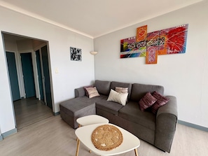 Una sala de estar moderna con un sofá seccional gris, una mesa de centro blanca con un posavasos tejido y un cuadro abstracto colorido en la pared.