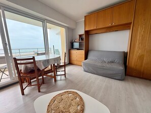 Ein Zimmer mit Balkon, einem Esstisch, einem grauen Sofa, Holzschränken und einem Fernseher.