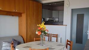 A dining area with a table set for four, a vase with flowers, wooden chairs, a sofa, and a kitchen in the background. - Comfortable Studio for 3 in Saint Jean de Monts, Near Beach (Saint-Jean-de-Monts)