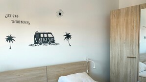 A bedroom with a bed, white pillows, a wooden headboard, a wall decal with a vintage surf truck and palm trees, and a wooden wardrobe. - Spacious apartment for 4, 200m from the beach, private parking included (Saint-Hilaire-de-Riez)