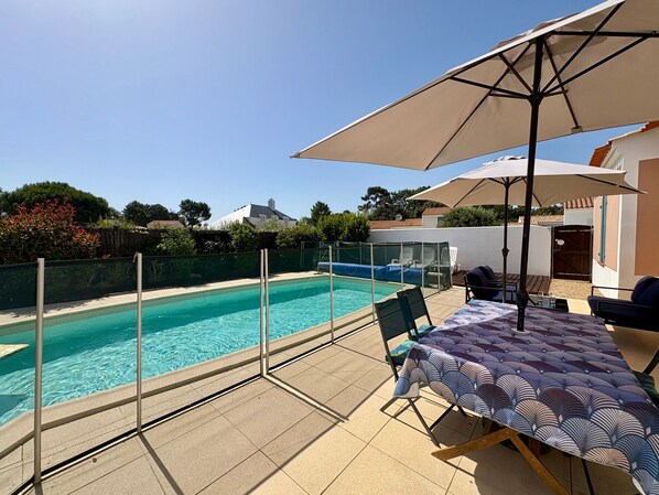 A swimming pool with a glass barrier, a covered outdoor seating area with a table and chairs, and a view of the surrounding landscape.