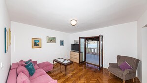 A living room with a pink sofa, a grey armchair, a wooden coffee table, and a TV on a wooden cabinet. There are three framed pictures on the wall, and a balcony with a view of the sea. - 4 bedroom nice home in Okrug Donji (Okrug Donji)