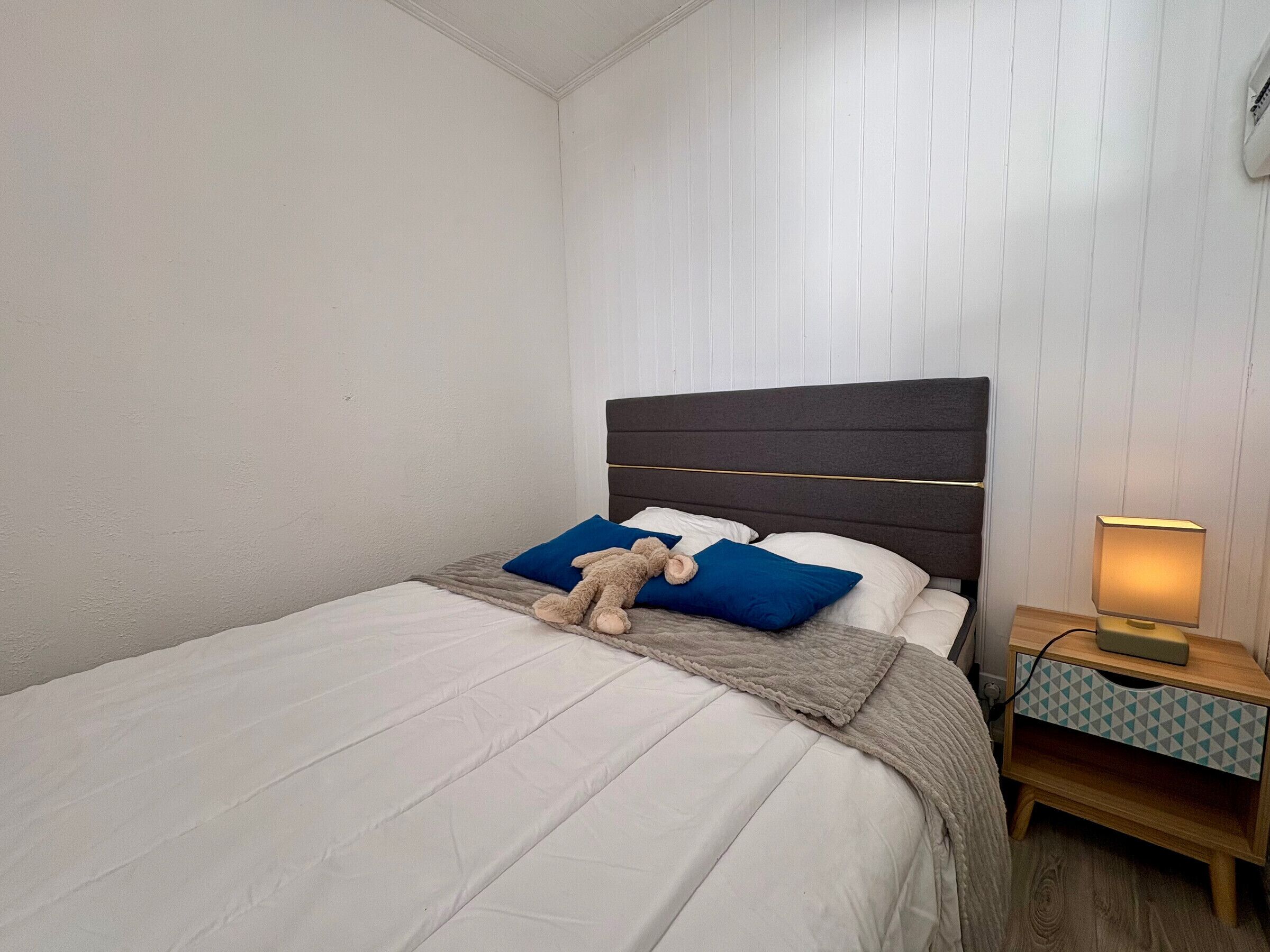 A neatly made bed with a brown teddy bear, blue pillows, and a grey blanket.