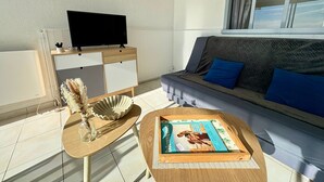 A modern living room with a sofa, a coffee table with a magazine, a TV on a stand, and a window with curtains. - Bright studio with alcove, sea-view balcony, private parking (Saint-Jean-de-Monts)