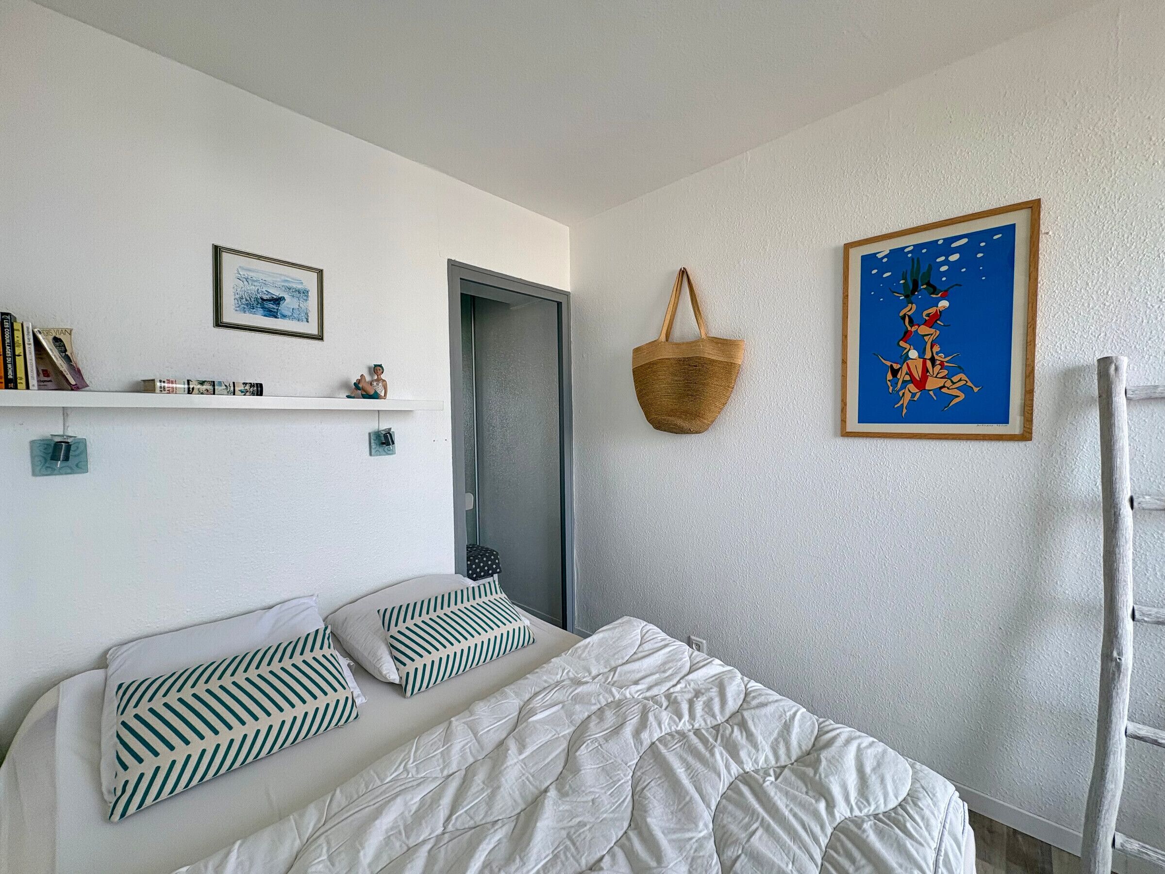Ein Schlafzimmer mit einem Bett, einem Wandregal mit Büchern und einer Figur, einer Tür, einer hängenden Ampel und einem gerahmten Bild.
