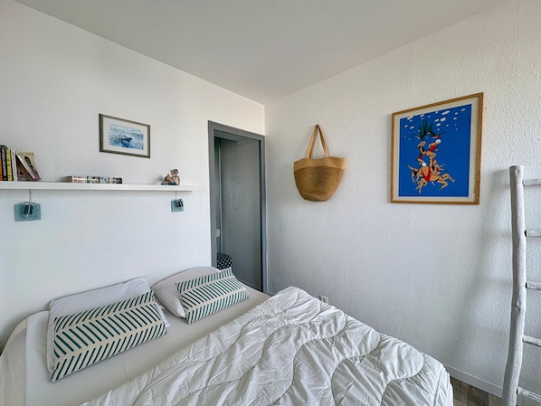 A bedroom with a bed, a wall-mounted shelf with books and a figurine, a door, a hanging basket, and a framed picture.