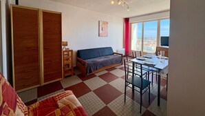 Una habitación con sofá, una mesa de comedor preparada para dos personas, un ventanal con vistas y un armario de madera.