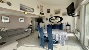 Ein Wohnzimmer mit einer Couch, einem Esstisch mit Stühlen, einem an der Wand montierten Fernseher und verschiedenen gerahmten Bildern an den Wänden.