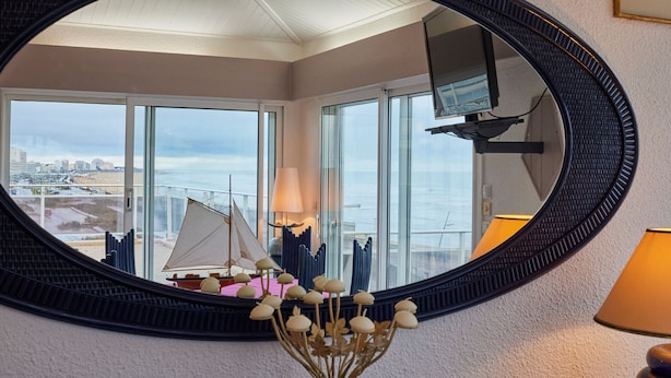 Ein Zimmer mit einem großen, ovalen Spiegel, einer kleinen Zimmerpflanze und Blick auf ein Segelboot durch große Fenster.