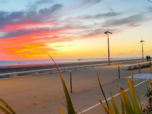 Une vue sur le coucher de soleil depuis une promenade en bord de mer, avec une barrière en béton séparant le chemin piétonnier de la plage.