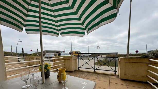 A terrace with a table set for dining, a striped umbrella, and a view of the parking area.