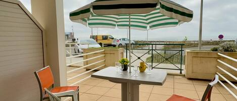 A balcony with a table and chairs, a striped umbrella, and a view of the sea and beach.