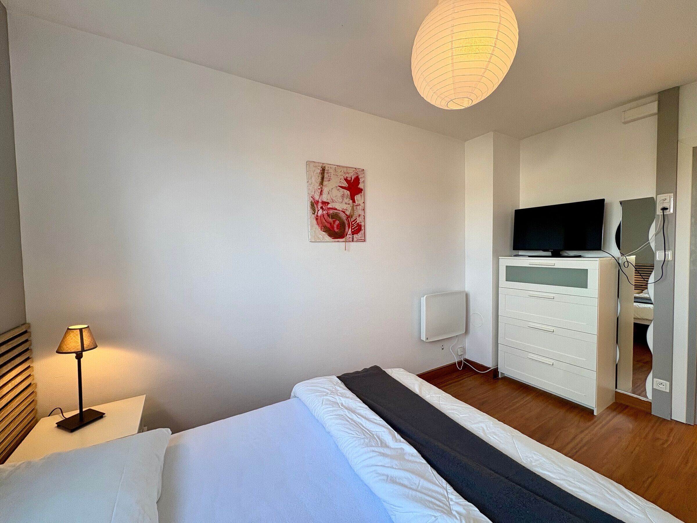Ein Hotelzimmer mit Bett, Nachttischlampe, Flachbildfernseher, Kommode und Wandheizung.