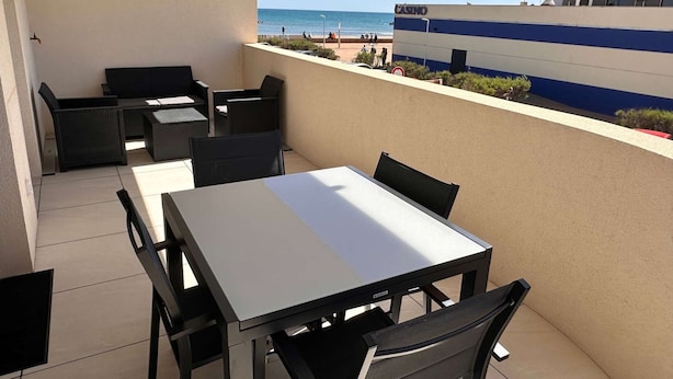 A balcony with a table and chairs, overlooking a beach and a casino building. - Beautiful T2 with large terrace and parking, near the sea (Valras-Plage)