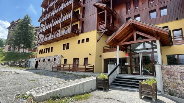 A multi-story wooden building with balconies, a stone wall, and a staircase leading to the entrance.
