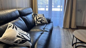 A black leather sofa with patterned cushions, a small wooden table, and a view of a wooden deck through sheer curtains.