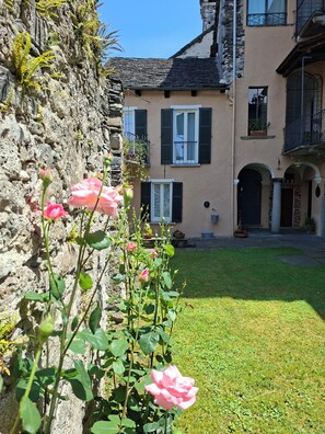 Un mur de pierres, agrémenté de fougères et d’un rosier au premier plan, un bâtiment à la façade en pierre et doté d’un balcon, et une cour verdoyante.