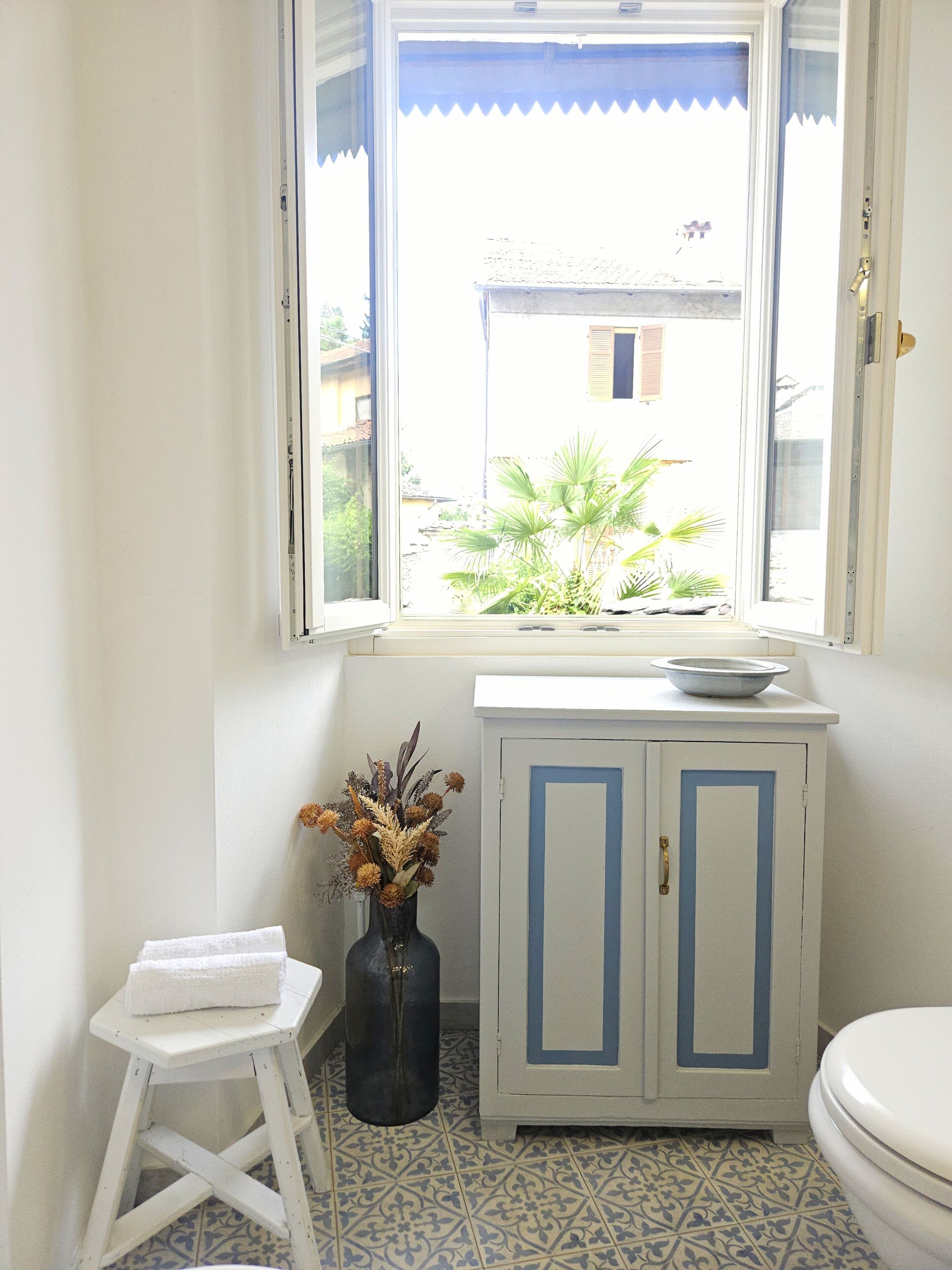 A bathroom with a window, a white stool, a cabinet, and a toilet.