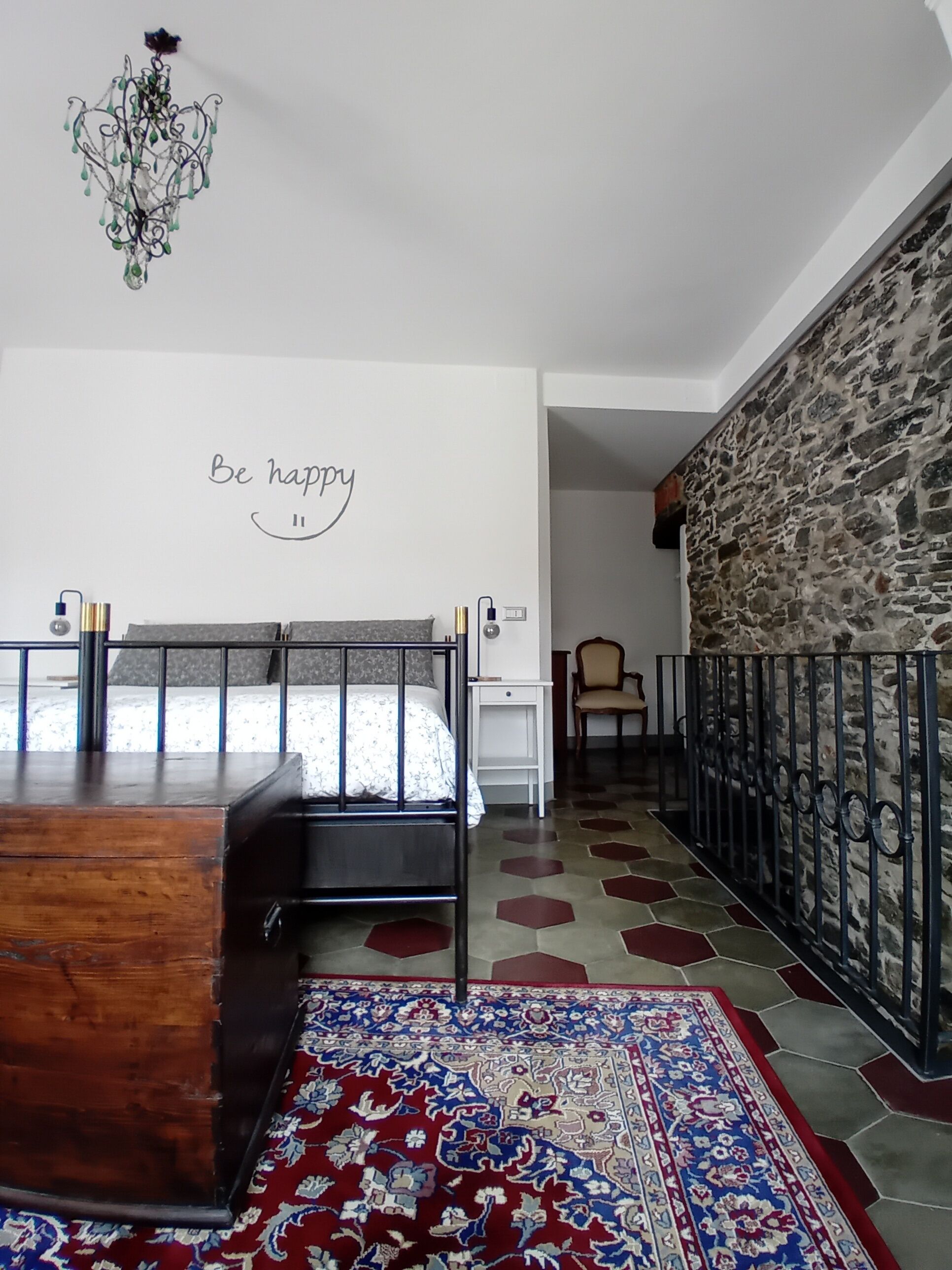 Ein Schlafzimmer mit einem hölzernen Bett, einem Teppich mit blau-rotem Muster, einer Steinwand und einem Schild mit der Aufschrift 'Be happy'.