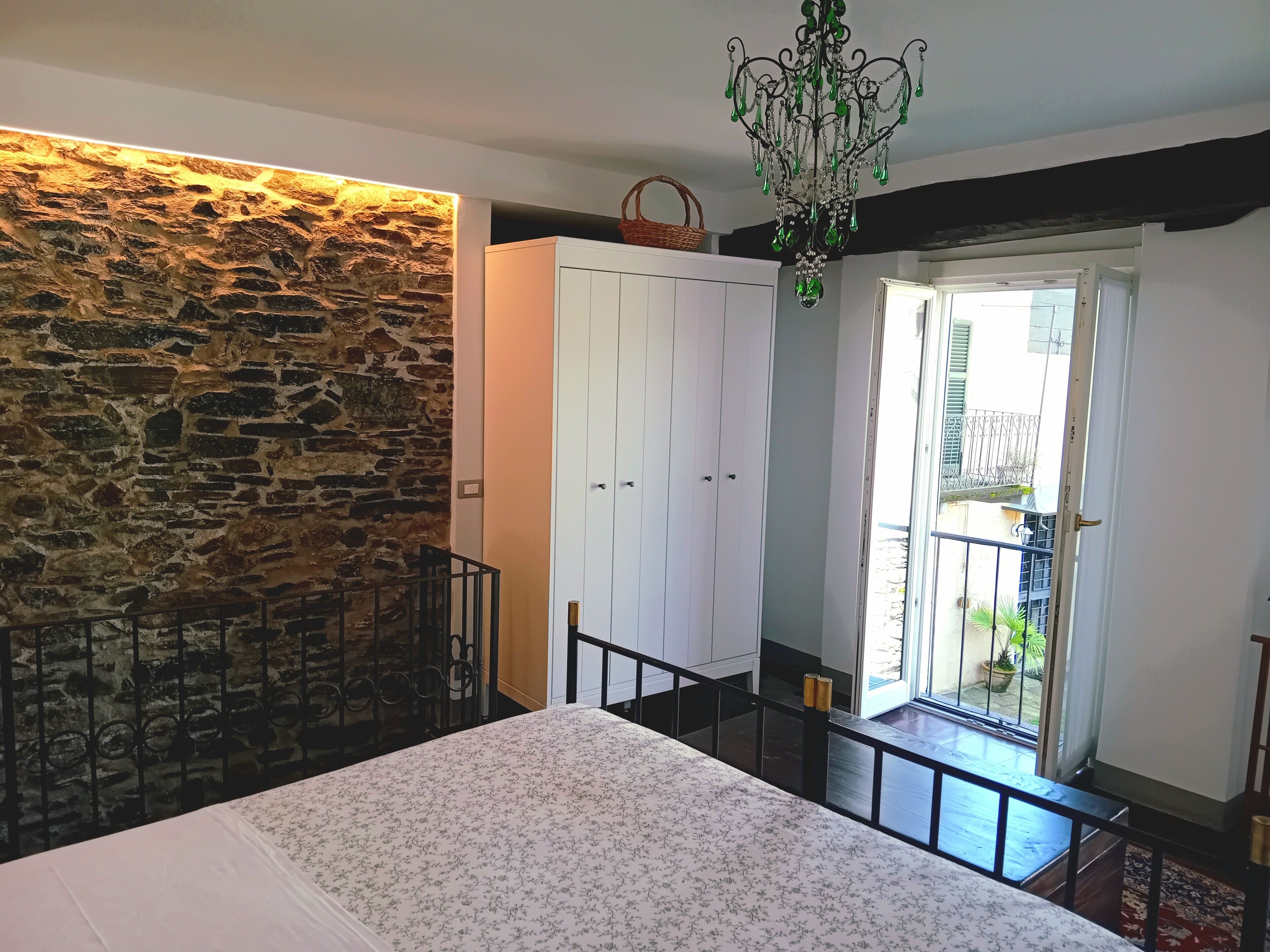 A bedroom with a stone wall, a white wardrobe, a bed with a floral bedspread, and a balcony with a view of the outside.