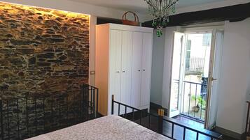 A bedroom with a stone wall, a white wardrobe, a bed with a floral bedspread, and a balcony with a view of the outside.