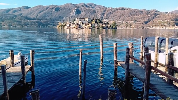 A lake with a wooden dock and a boat. In the background, there is a town with buildings on a hill.