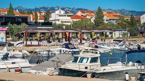 A marina with boats docked, a covered seating area, and buildings with red roofs in the background. - Deluxe Villa Laura With Pool Studio 1b (Novalja)